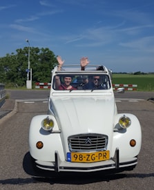 A white vintage car, specifically a Citroën 2CV, is pictured on a road with two people sitting inside. The person on the left is wearing a red top and has both hands raised. The person on the right appears to be the driver and is wearing sunglasses. The license plate of the car reads 98-ZG-PR with a blue NL symbol indicating it's from the Netherlands. There is a clear sky with some light clouds and greenery in the background.