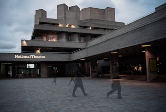 An architectural structure with a modernist design, featuring a large concrete building labeled 'National Theatre'. The building has multiple levels with stark geometric shapes and is illuminated by exterior lights. In the foreground, blurred figures of people are walking across a paved area, creating a sense of motion and life against the static backdrop of the theatre. The sky appears overcast, adding to the somber ambiance.
