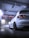 A sleek white car is parked inside a modern car wash facility. The lighting is dramatic, with a cool, bluish hue reflecting on the wet floor. The car's black rims and taillights are prominently visible, adding to its sporty appearance.