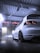 A sleek white car is parked inside a modern car wash facility. The lighting is dramatic, with a cool, bluish hue reflecting on the wet floor. The car's black rims and taillights are prominently visible, adding to its sporty appearance.