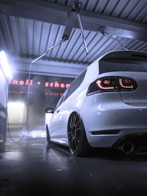 A sleek white car is parked inside a modern car wash facility. The lighting is dramatic, with a cool, bluish hue reflecting on the wet floor. The car's black rims and taillights are prominently visible, adding to its sporty appearance.