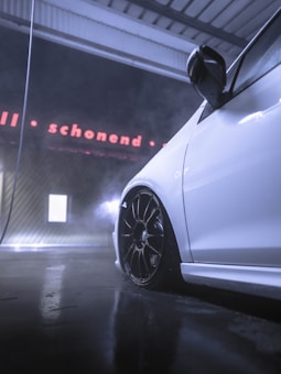 A white car is parked indoors with one side visible, showing a sleek design and black wheels. The flooring reflects a metallic sheen, and there is mist or vapor around. A sign in the background reads 'schonend' in red neon letters with a modern industrial setting.