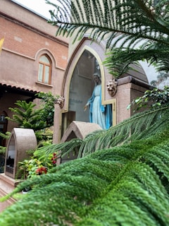 A religious statue of a figure in blue and white robes is enclosed within a shrine with a pointed arch and glass front. The scene is surrounded by greenery, including lush ferns and flowering plants. The building in the background has arched windows and is made of reddish-brown brick.
