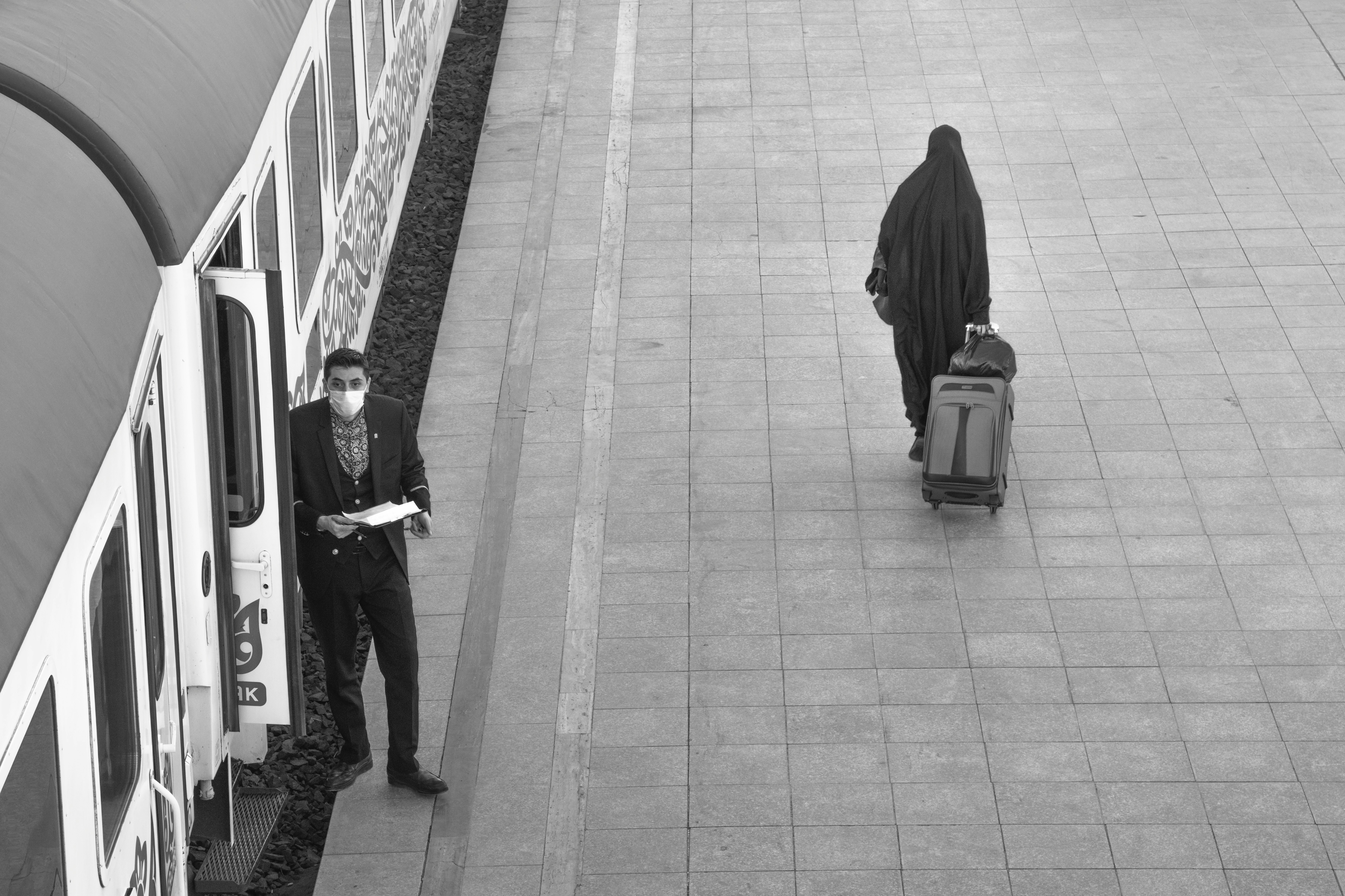grayscale photo of woman in black hijab standing beside train