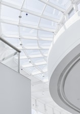 A modern architectural interior featuring a large, translucent glass ceiling allowing natural light to flood the space. The curved and straight lines of the ceiling and walls create an open and airy environment. Minimalist design elements and neutral colors emphasize the contemporary style.