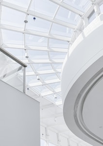 A modern architectural interior featuring a large, translucent glass ceiling allowing natural light to flood the space. The curved and straight lines of the ceiling and walls create an open and airy environment. Minimalist design elements and neutral colors emphasize the contemporary style.