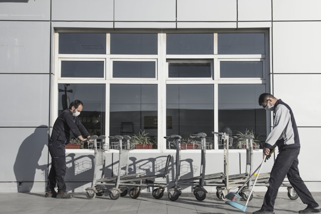 Two people wearing masks are engaged in cleaning activities outside a building with large windows. One person is pushing a line of shopping carts, while the other is sweeping the ground with a broom. The background consists of a paneled wall and a window with visible reflections.