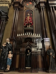 The image features a detailed wooden altar with religious statues. At the top center, a statue dressed in an ornate red robe is displayed, surrounded by an intricate arch design. Below, smaller statues are placed, including a figure holding a child on the left and another figure in a religious habit on the right. The woodwork is dark and polished, with elaborate carvings and decorative elements.