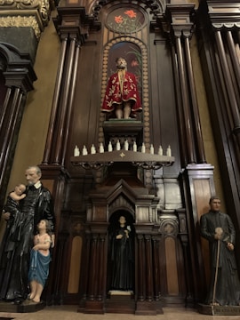 The image features a detailed wooden altar with religious statues. At the top center, a statue dressed in an ornate red robe is displayed, surrounded by an intricate arch design. Below, smaller statues are placed, including a figure holding a child on the left and another figure in a religious habit on the right. The woodwork is dark and polished, with elaborate carvings and decorative elements.