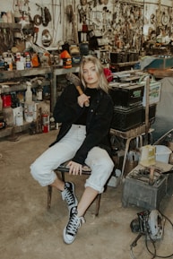 A person is sitting on a chair in a cluttered workshop filled with various tools and machinery. The individual is holding a tool, likely a hammer, and is wearing casual clothing including a black jacket and sneakers. The background features shelves stacked with metallic parts, cans, and mechanical equipment, contributing to an industrial and rustic atmosphere.