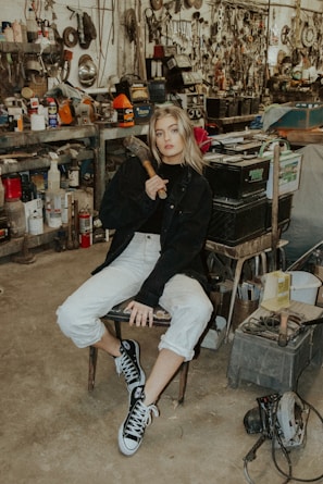 A person is sitting on a chair in a cluttered workshop filled with various tools and machinery. The individual is holding a tool, likely a hammer, and is wearing casual clothing including a black jacket and sneakers. The background features shelves stacked with metallic parts, cans, and mechanical equipment, contributing to an industrial and rustic atmosphere.