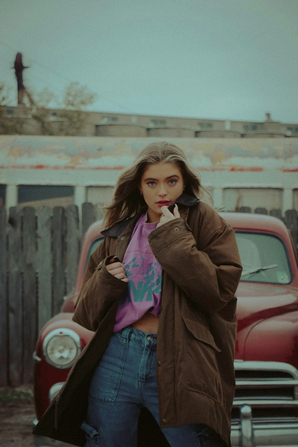 woman in brown coat and blue denim jeans standing beside red car during daytime