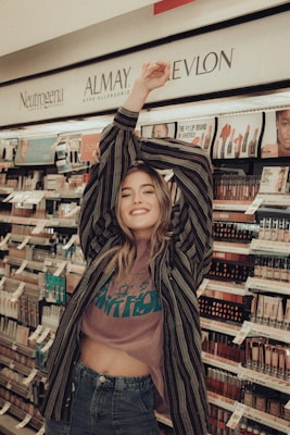 A person smiling and posing in front of shelves filled with cosmetics and beauty products, wearing a striped shirt and a casual outfit.