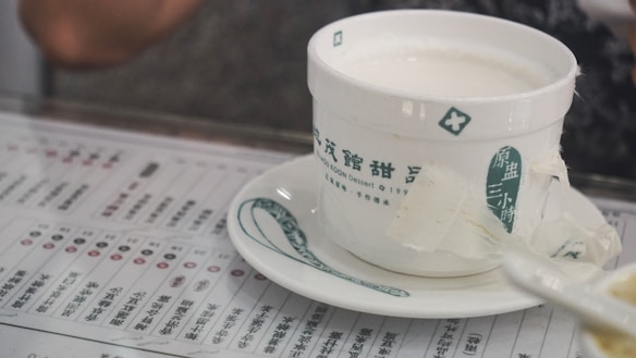 A white ceramic bowl with green Chinese characters on the side rests on a matching saucer. It is filled with a creamy white liquid, possibly a dessert. The bowl is placed on top of a menu with various text and symbols printed in Chinese.