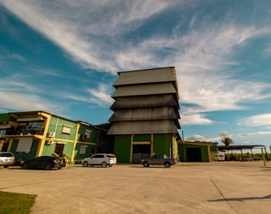 An industrial building complex with a large, tiered structure in the center, surrounded by smaller buildings of similar architectural style. The scene includes parked cars on a wide concrete area, with a partly cloudy sky and palm trees in the background.