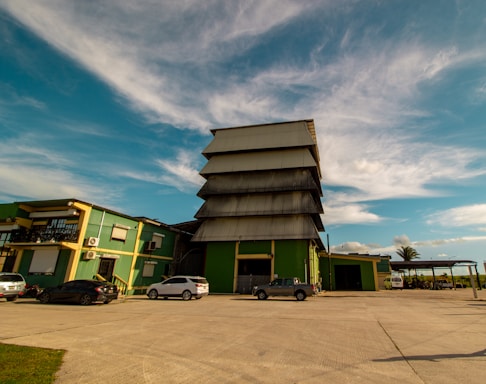 An industrial building complex with a large, tiered structure in the center, surrounded by smaller buildings of similar architectural style. The scene includes parked cars on a wide concrete area, with a partly cloudy sky and palm trees in the background.