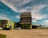 An industrial building complex with a large, tiered structure in the center, surrounded by smaller buildings of similar architectural style. The scene includes parked cars on a wide concrete area, with a partly cloudy sky and palm trees in the background.