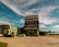An industrial building complex with a large, tiered structure in the center, surrounded by smaller buildings of similar architectural style. The scene includes parked cars on a wide concrete area, with a partly cloudy sky and palm trees in the background.