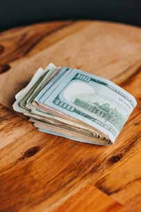 A stack of various denominations of paper currency, including prominently visible 100-dollar bills, placed on a wooden surface.