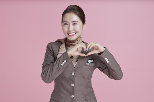 A person in a brown uniform with a scarf stands against a pink background, making a heart shape with her hands and smiling warmly.