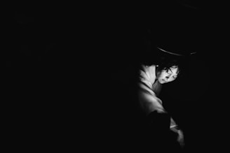 A black and white photograph highlighting a person's face partially illuminated against a dark background. The lighting creates a dramatic effect, emphasizing the contours of the face and arm, with a focus on expression and emotion.
