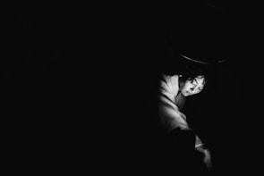 A black and white photograph highlighting a person's face partially illuminated against a dark background. The lighting creates a dramatic effect, emphasizing the contours of the face and arm, with a focus on expression and emotion.