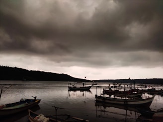 Dark, ominous clouds loom over a body of water where several small boats are docked. The water is calm, reflecting the boats and the cloudy sky. The distant shoreline is slightly visible, with some boats seemingly anchored farther from the shore. The mood is somber and tranquil.