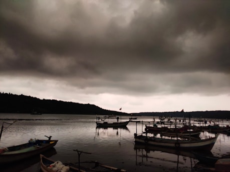 Dark, ominous clouds loom over a body of water where several small boats are docked. The water is calm, reflecting the boats and the cloudy sky. The distant shoreline is slightly visible, with some boats seemingly anchored farther from the shore. The mood is somber and tranquil.