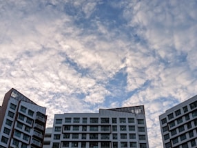 A cluster of modern multi-story buildings with a white and brown design contrasts against a sky filled with fluffy, scattered clouds.