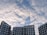 A cluster of modern multi-story buildings with a white and brown design contrasts against a sky filled with fluffy, scattered clouds.