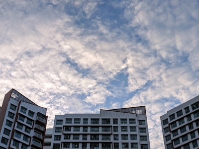 A cluster of modern multi-story buildings with a white and brown design contrasts against a sky filled with fluffy, scattered clouds.