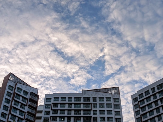 A cluster of modern multi-story buildings with a white and brown design contrasts against a sky filled with fluffy, scattered clouds.