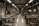 A textile workshop with numerous sewing machines arranged in two rows along a spacious, well-lit room. Several workers are seated at the machines, diligently crafting or sewing garments. The space is organized yet filled with fabric pieces scattered on the floor, contributing to a busy atmosphere. Overhead, fluorescent lights evenly illuminate the area, and large windows line the walls, allowing natural light to filter in.