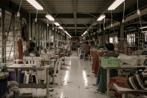 A textile workshop with numerous sewing machines arranged in two rows along a spacious, well-lit room. Several workers are seated at the machines, diligently crafting or sewing garments. The space is organized yet filled with fabric pieces scattered on the floor, contributing to a busy atmosphere. Overhead, fluorescent lights evenly illuminate the area, and large windows line the walls, allowing natural light to filter in.