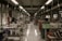 A textile workshop with numerous sewing machines arranged in two rows along a spacious, well-lit room. Several workers are seated at the machines, diligently crafting or sewing garments. The space is organized yet filled with fabric pieces scattered on the floor, contributing to a busy atmosphere. Overhead, fluorescent lights evenly illuminate the area, and large windows line the walls, allowing natural light to filter in.