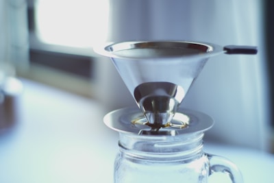 A metallic coffee filter rests atop a glass jar, positioned indoors with diffused natural light streaming in from a nearby window. The filter has a conical shape and is designed for pour-over coffee brewing.