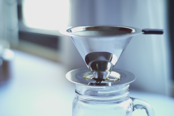 A metallic coffee filter rests atop a glass jar, positioned indoors with diffused natural light streaming in from a nearby window. The filter has a conical shape and is designed for pour-over coffee brewing.
