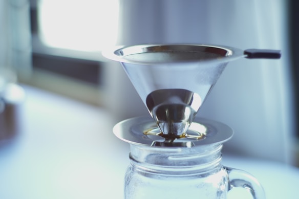 A metallic coffee filter rests atop a glass jar, positioned indoors with diffused natural light streaming in from a nearby window. The filter has a conical shape and is designed for pour-over coffee brewing.