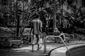 A black and white photograph shows a man walking along a paved path in a garden, carrying a bottle and a bag. In the background, another person sits on a bench surrounded by plants, seemingly engrossed in reading. The area is lush with tall trees and foliage, creating a serene outdoor setting.