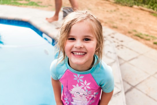 A young child with a big smile, wearing a colorful swimsuit with a character design, stands next to a pool. The background features a poolside area with some grass and paving stones.