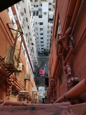 An aerial view down a narrow alleyway bordered by two tall, closely situated apartment buildings. The walls of the buildings are lined with numerous pipes, air conditioning units, and external balconies. Below, a small courtyard is visible with a single red tarp-covered structure and various scattered items.