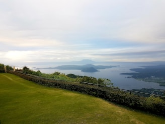 A scenic landscape features a lush green lawn in the foreground, bordered by trees and shrubbery. In the distance, a serene body of water stretches out toward a mountain range under an expansive sky with scattered clouds.