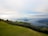 A scenic landscape features a lush green lawn in the foreground, bordered by trees and shrubbery. In the distance, a serene body of water stretches out toward a mountain range under an expansive sky with scattered clouds.