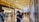A modern shopping mall corridor with glossy floors and a high, illuminated ceiling. Various high-end brand stores, including Dior and Louis Vuitton, are visible. Shoppers, some wearing masks, are walking and browsing their phones.