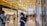 A modern shopping mall corridor with glossy floors and a high, illuminated ceiling. Various high-end brand stores, including Dior and Louis Vuitton, are visible. Shoppers, some wearing masks, are walking and browsing their phones.