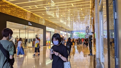 A modern shopping mall corridor with glossy floors and a high, illuminated ceiling. Various high-end brand stores, including Dior and Louis Vuitton, are visible. Shoppers, some wearing masks, are walking and browsing their phones.