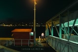 A dimly lit dock scene at night featuring a red structure labeled 'ZAGAIA-LX-157-AL' and illuminated by a streetlamp. A person stands near the edge of the dock beside a metallic structure with teal lighting. Across the water, a cityscape is visible with scattered lights in the distance, implying an urban environment.