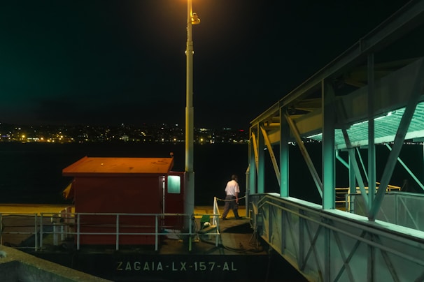 A dimly lit dock scene at night featuring a red structure labeled 'ZAGAIA-LX-157-AL' and illuminated by a streetlamp. A person stands near the edge of the dock beside a metallic structure with teal lighting. Across the water, a cityscape is visible with scattered lights in the distance, implying an urban environment.
