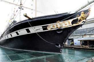 A large black and white ship with intricate golden decorations on the bow, situated in a dry dock. The ship has sails and masts adorned with colorful flags, and there are industrial buildings in the background.