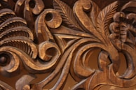Intricate wooden carvings display detailed scrollwork and leaf motifs with smooth, flowing lines, highlighted by the natural grain and texture of the wood.